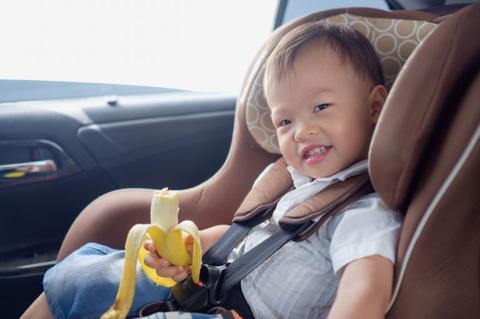 Cute little Asian 18 months / 1 year old toddler baby boy child sitting in safety carseat holding & enjoy eating banana, Happy traveling with child concept, Little Traveler safety, road trip concept