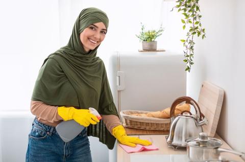 Domestic Chores. Smiling happy arab woman in headscarf and protecitve yellow rubber gloves cleaning countertop standing at kitchen, holding spray bottle and microfiber cloth, looking at camera