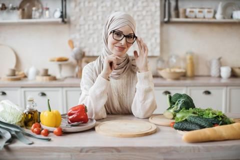 Young smiling muslim woman in hijab sitting at table with fresh vegetables and baguette in modern light kitchen, preparing to make a diet salad from fresh vegetarian ingredients.